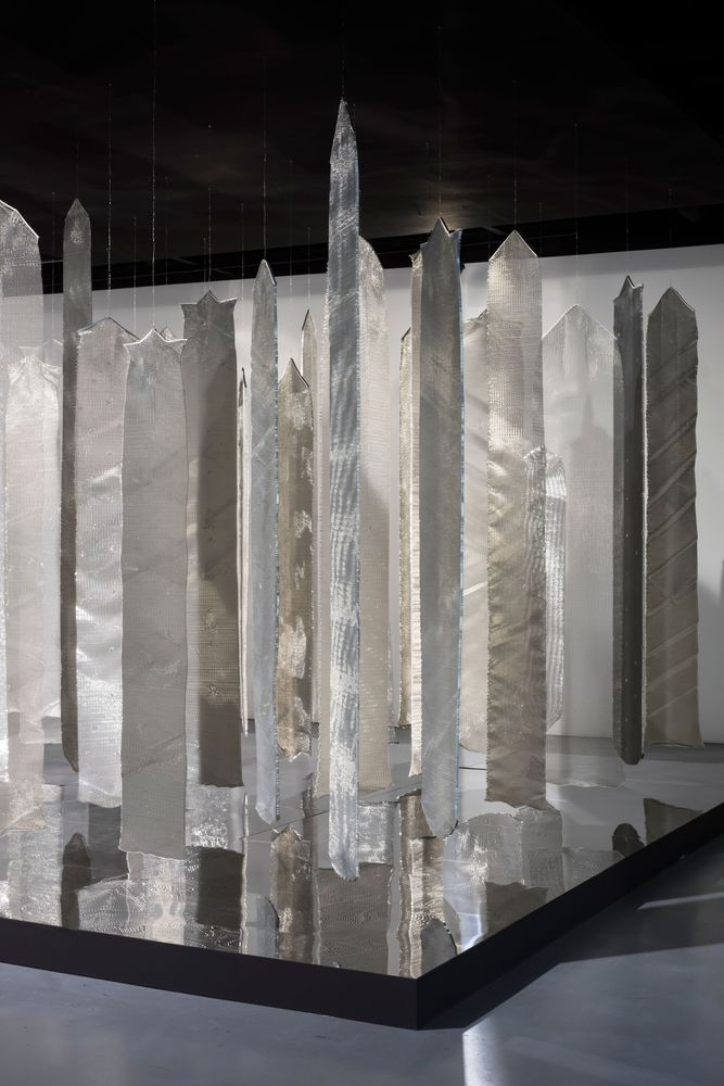 Eine Installation aus mehreren metallisch glänzenden Stoffen hängt von der Decke des Raumes. 