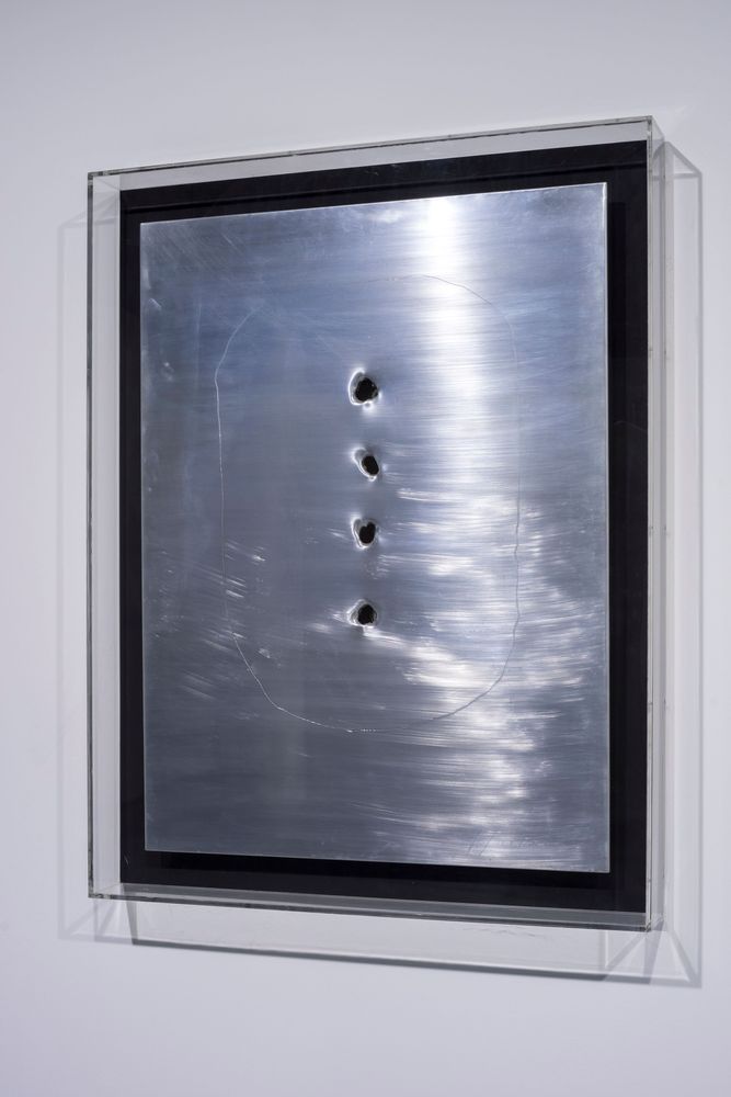 Eine metallische Fläche, in die vier Löcher gebohrt wurden, hängt in einem Glaskasten.