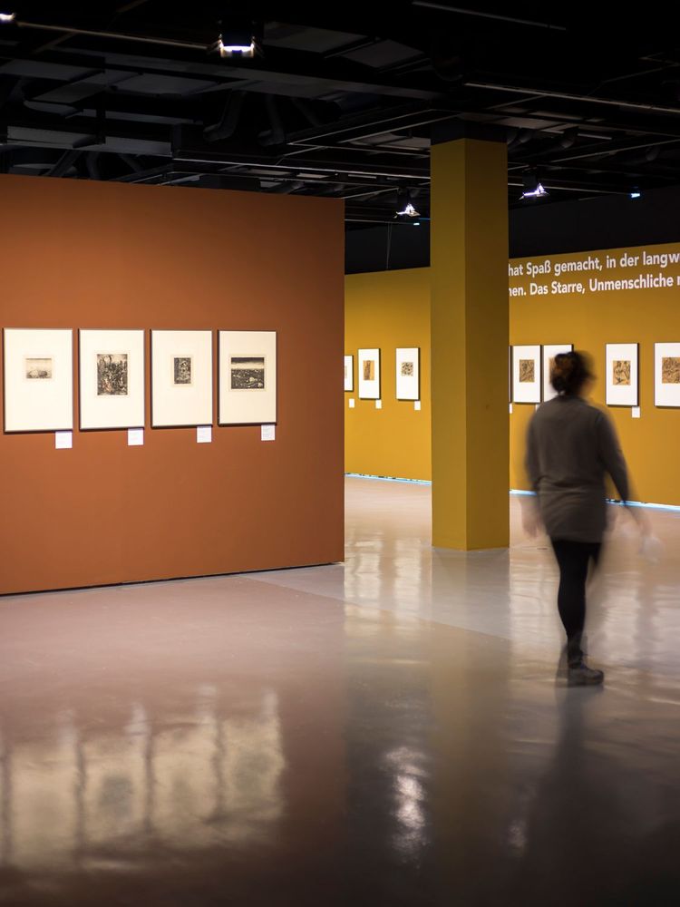 An einer braunen und an einer gelb-braunen Wand hängen verschiedene Bilder, rechts sieht man die verschwommene Silhouette einer sich bewegenden Frau.