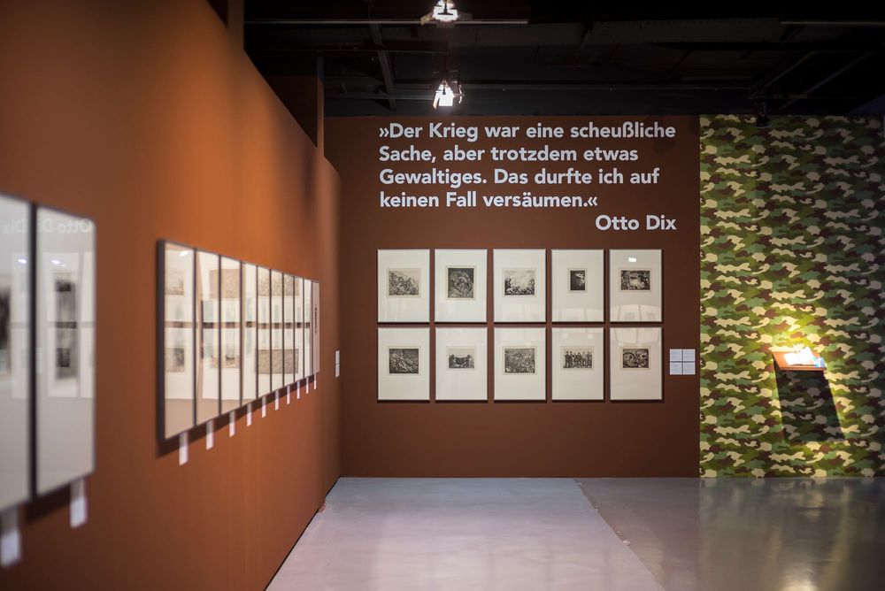Die braunen Wände auf der linken und hinteren Seite des Bildes sind mit verschiedenen Bildern behangen. Ein teil der wand auf der rechten Seite ist in Tarnfarben gemustert.