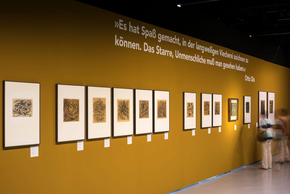 An einer braun-gelben Wand hängen verschiedene Bilder in einer Reihe. Die Silhouetten von drei Frauen am rechten Bildrand betrachten die Bilder. 