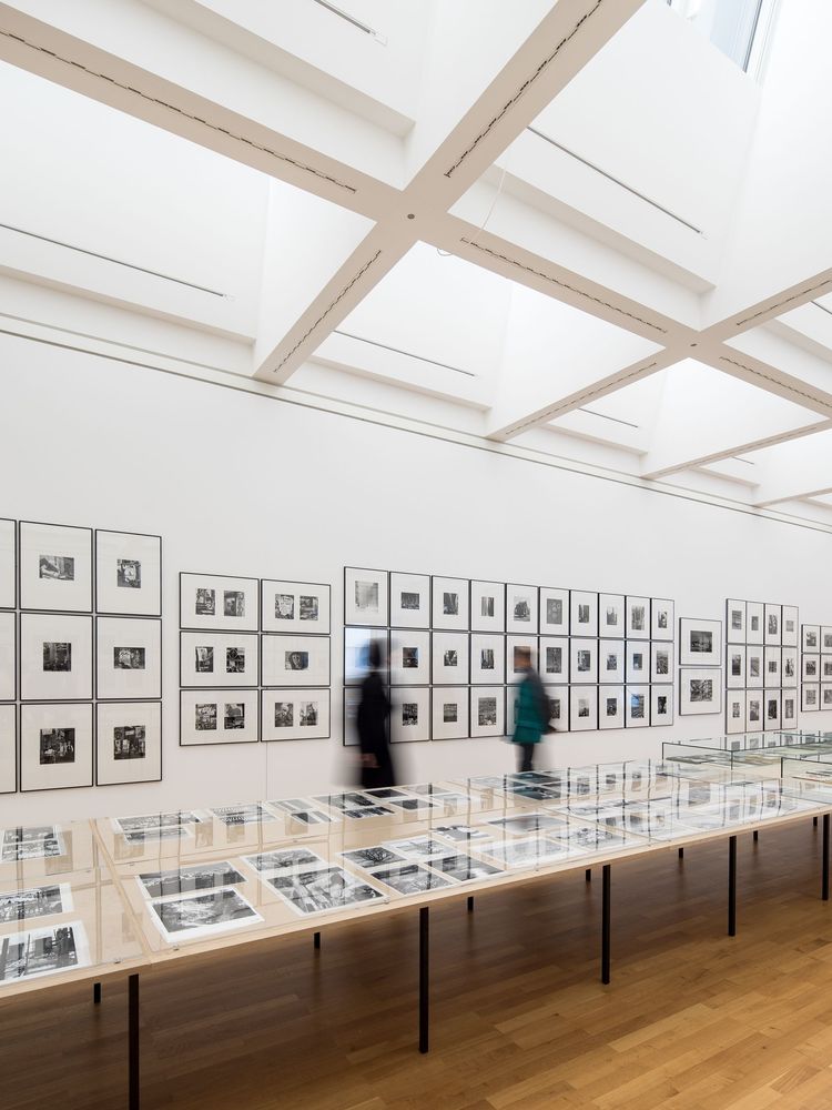 Die Ausstellungansicht zeigt eine Reihe von mehreren Tischen auf denen Fotografien verteilt sind, an den hinteren Wänden sind ebenfalls sehr viele Fotografien aufgehängt. Vor der wand erkennt man die Silhouetten von zwei sich bewegenden Personen.