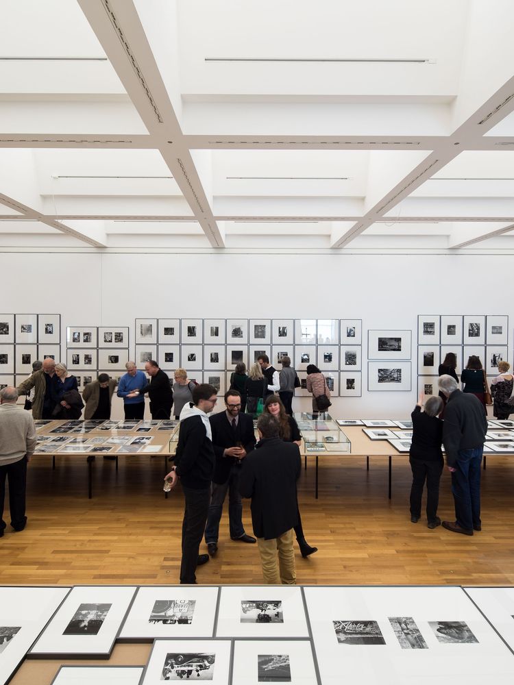 Die Ausstellungansicht zeigt zwei Reihen von mehreren Tischen auf denen Fotografien verteilt sind. An der hinteren Wand sind ebenfalls sehr viele Fotografien aufgehängt. Um die Tische herum bewegen oder unterhalten sich Besucher*innen.