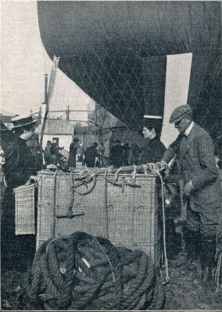 Die historische Aufnahme zeigt einen Mann der neben einem Ballonkorb steht und etwas in diesen legt. Dahinter stehen zwei Frauen sich am Korb gegenüber und unterhalten sich. 