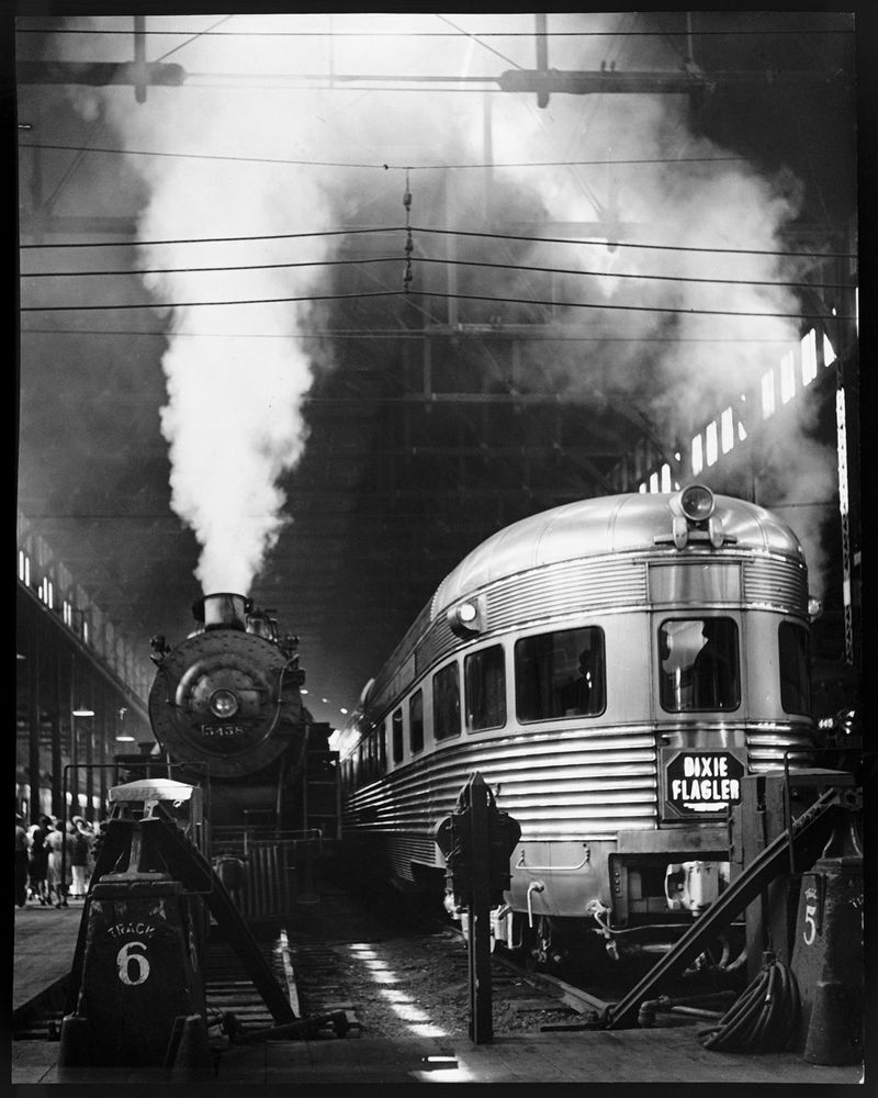 Die schwarz-weiß Fotografie zeigt eine ältere Bahnstation. Neben einer dampfenden Lokomotive steht ein großer Wagon.