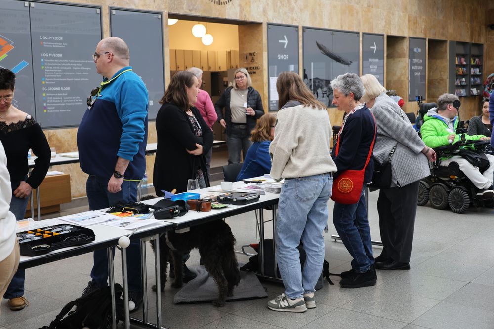 Der Markt der Möglichkeiten im Foyer zeigt Hilfsmittel und Brillen, um Augenkrankheiten zu simulieren auf langen Tischen. Um die Tische stehen einige Menschen, die im Gespräch sind. 