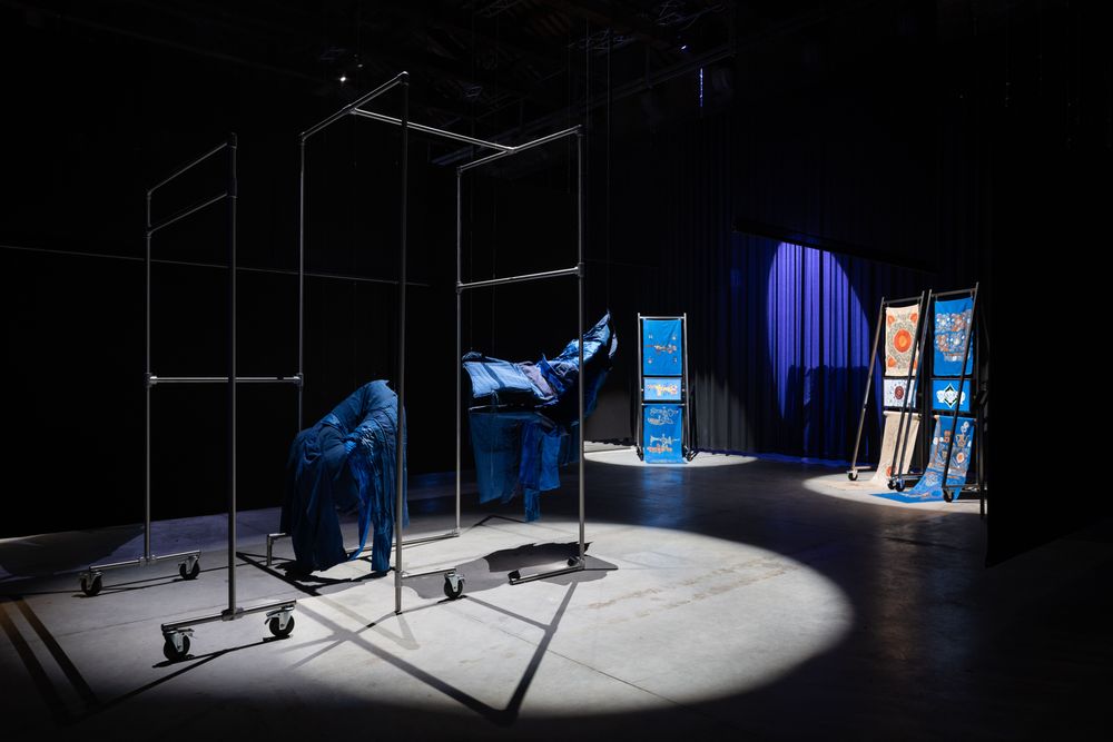 Installationsansicht in einem abgedunkelten Raum. Im Vordergrund stehen leere Metallgestelle auf Rollen, teils mit blauen Stoffen oder Planen überzogen und in einem Lichtkegel inszeniert.