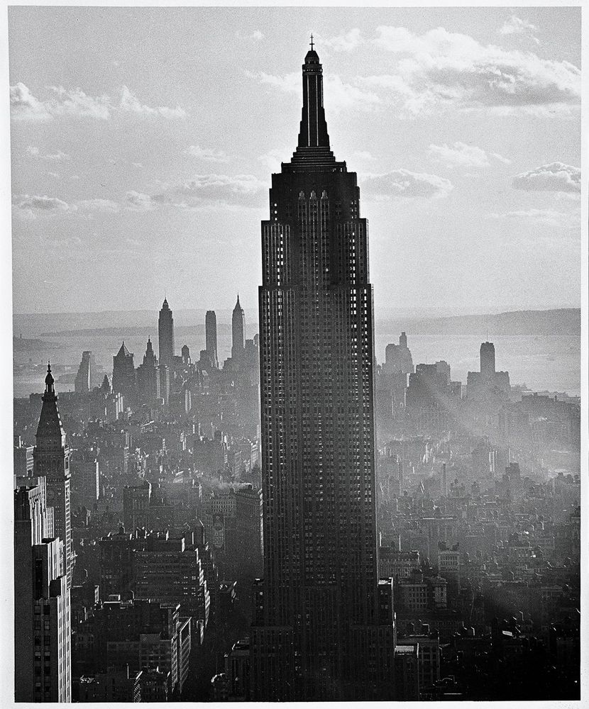 Die schwarz-weiß Fotografie zeigt das Empire State Building in New York. Im Hintergrund erstrecken sich weitere Hochhäuser.