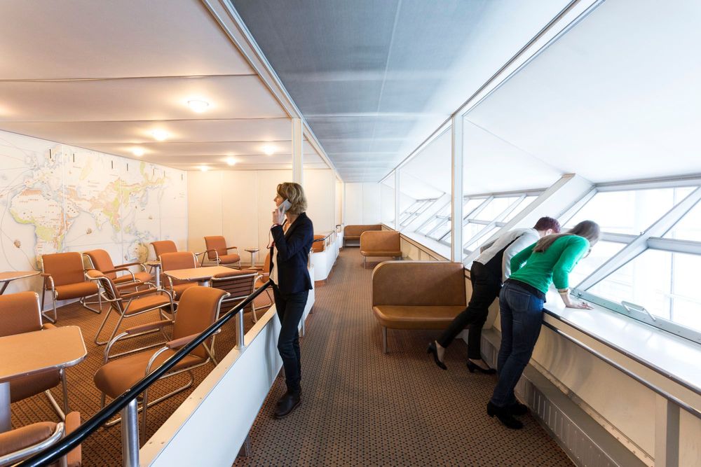 Farbfoto der Hindenburg-Teilrekonstruktion mit Blick ins Promenadendeck. Zwei Besucherinnen schauen rechts aus den Panoramafenstern. Eine Frau steht links und betrachtet die an die Wand gesprühte bunte Weltkarte mit einem Audioguide am Ohr.
