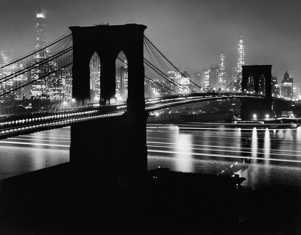 Die schwarz-weiß Fotografie zeigt die Silhouette einer riesigen Brücke bei Nacht, der Brooklyn Bridge. Dahinter funkeln die Lichter der Hochhäuser von New York.