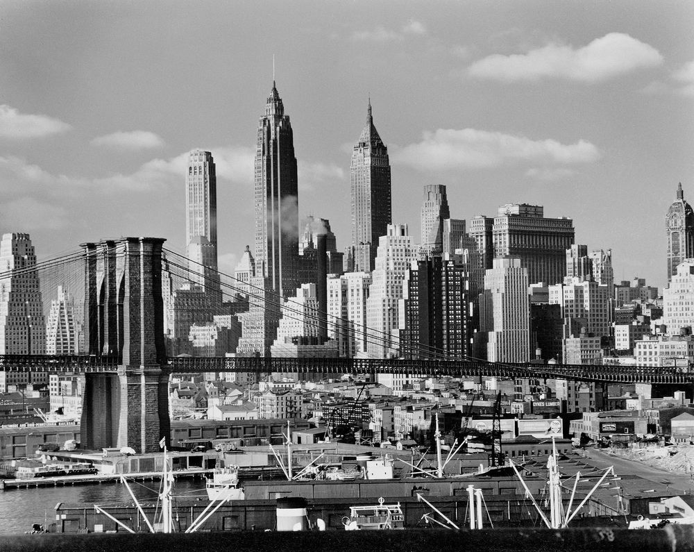Die schwarz-weiß Fotografie zeigt die riesige Brooklyn Bridge. Darunter sind Hafengebäude zu erkennen und dahinter erstrecken sich dutzende Hochhäuser, die die Skyline von Manhattan bilden.