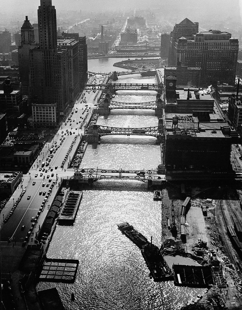 Die schwarz-weiß Fotografie zeigt einen Fluss der sich durch eine Stadt zieht. Kleine Brücken verbinden Straßen miteinander. Im Hintergrund und an den Rändern des Bildes sind viele kleine Gebäude und Hochhäuser zu erkennen.