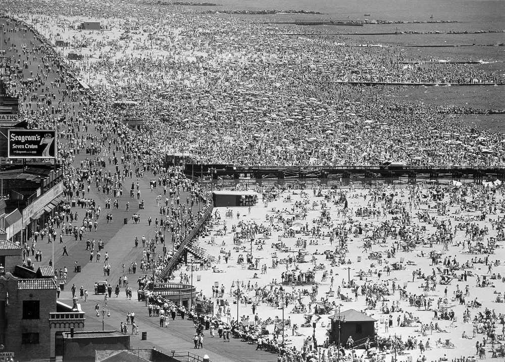 Die schwarz-weiß Fotografie zeigt einen völlig überfüllten Strand. Vor lauter Menschen und Sonnenschirmen sieht man den Sandstrand fast nicht mehr. Die Strandpromenade dahinter ist voll mit Fußgänger*innen.