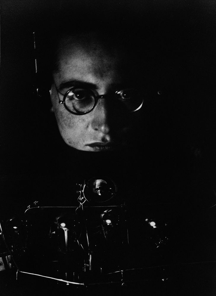 Die schwarz-weiß Fotografie zeigt den Künstler Andreas Feininger im Selbstporträt. Das Bild ist sehr dunkel und man erkennt fast nur seine rechte Gesichtshälfte. Er trägt eine runde Brille und unter seinem Gesicht ist eine Kamera zu erahnen. 