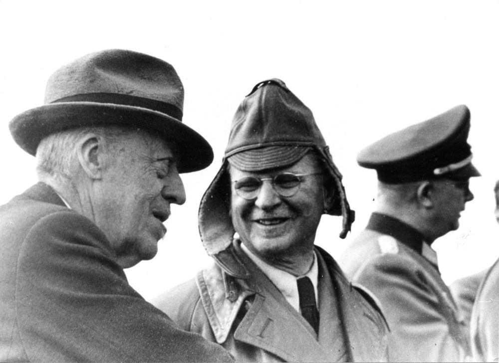 Hugo Eckener im angeregten Gespräch mit Karl Maybach. Eckener trägt Hut, Maybach eine Fliegermütze. Im Hintergrund ein Uniformierter Militärvertreter, dessen Gesicht jedoch abgewandt ist.