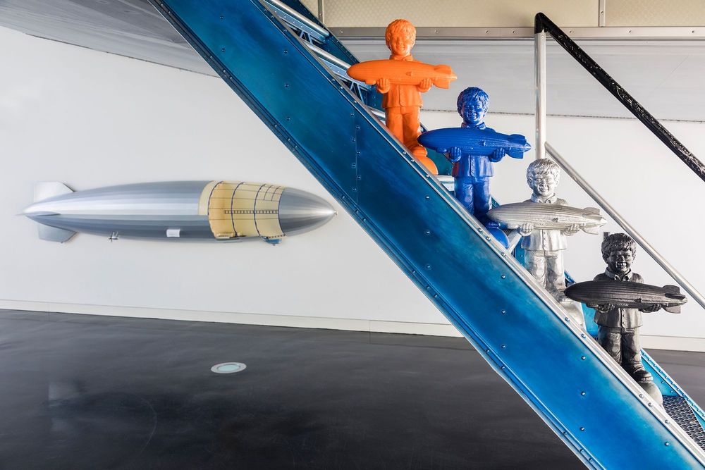 Auf den Treppenstufen eines Fallreeps stehen vier Zeppelin Jungen in den Farben orange, blau, silber und schwarz. Die Figuren zeigen einen Jungen, der einen Zeppelin in beiden Händen hält.