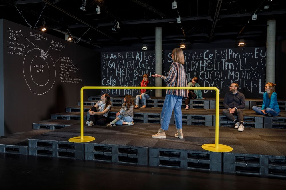 Ein moderner, dunkler Ausstellungsraum mit gestuften Sitzflächen aus Paletten. Mehrere Personen sitzen oder stehen, während eine Frau im Vordergrund hinter einer gelben Barriere steht und etwas zu erzählen scheint.