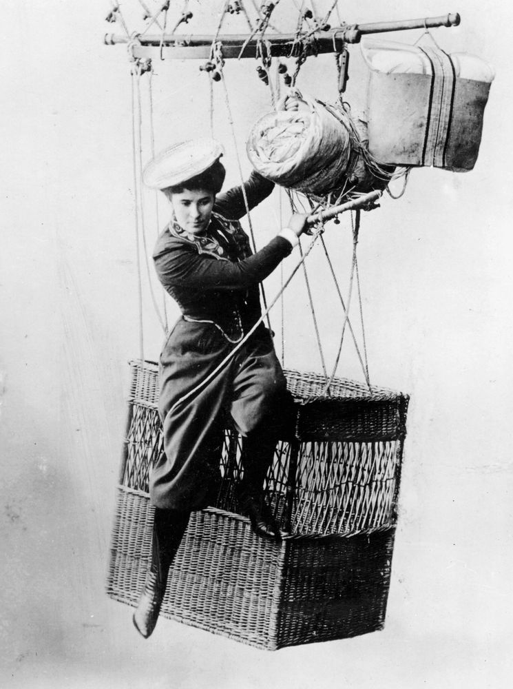 Die historische Aufnahme zeigt eine Luftakrobatin, die außen am Rand eines Ballonkorbes sitzt. Sie hält sich nur an Seilen fest und eines ihrer Beine hängt in der Luft. 