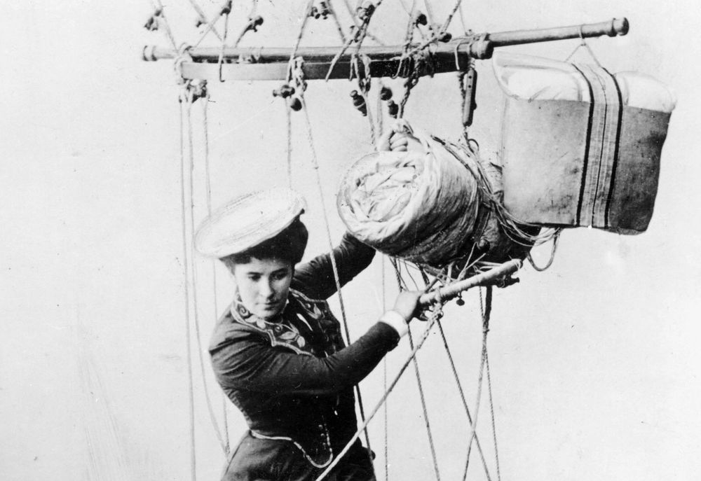 Die historische Aufnahme zeigt eine Luftakrobatin, die außen am Rand eines Ballonkorbes sitzt. Sie hält sich nur an Seilen fest und eines ihrer Beine hängt in der Luft. 