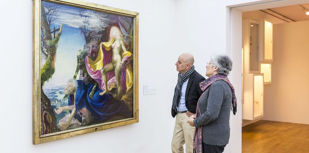 Zwei Personen betrachten ein großes, farbenfrohes Gemälde, das an einer weißen wand hängt. Das Bild zeigt eine Szene mit mehreren Figuren in einer Landschaft.
