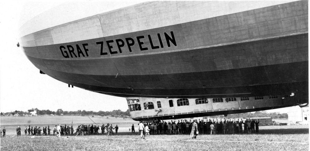 Die historische Aufnahme zeigt den Anschnitt eines Zeppelins und dessen Gondel, der von einer Haltemannschaft bestehend aus sehr vielen Männern zu Boden gezogen wird.