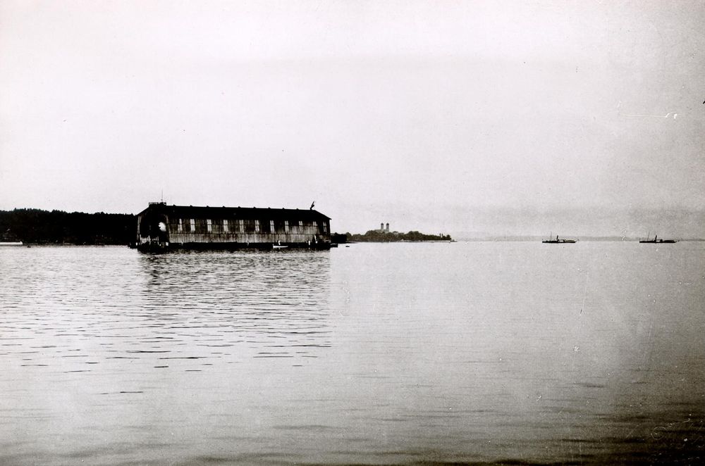 Blick auf die schwimmende Luftschiffhalle auf dem Bodensee bei Manzell. In der Halle ist der Bug des Luftschiffs sichtbar. Im Hintergrund stehen die Türme der Friedrichshafener Schlosskirche. Rechts im Hintergrund auf dem See fahren zwei Dampfer