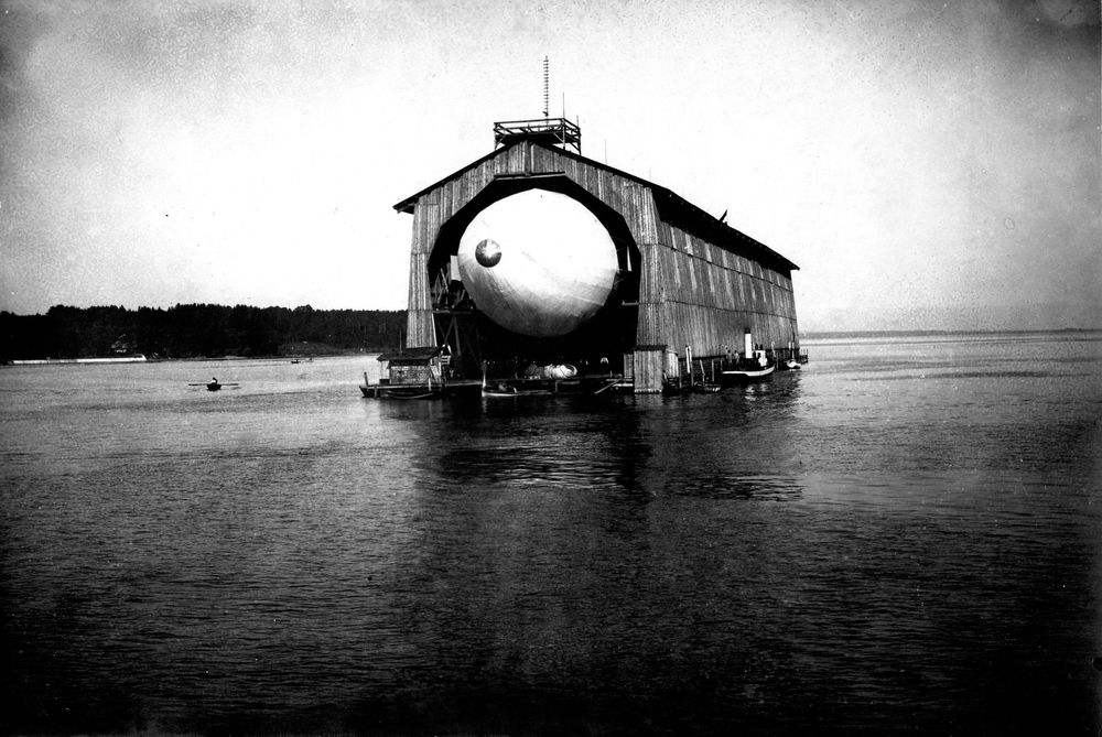 Der Zeppelin lugt aus der Halle hervor. Man sieht das Luftschiff frontal. An der Längsseite der Halle liegt ein Schiff.