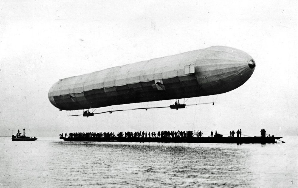 Der Zeppelin erhebt sich vom mit Menschen vollbesetzten Ponton. Zwei Gondeln hängen unter dem Luftschiff. Links vom Ponton befindet sich ein kleines Dampfboot.