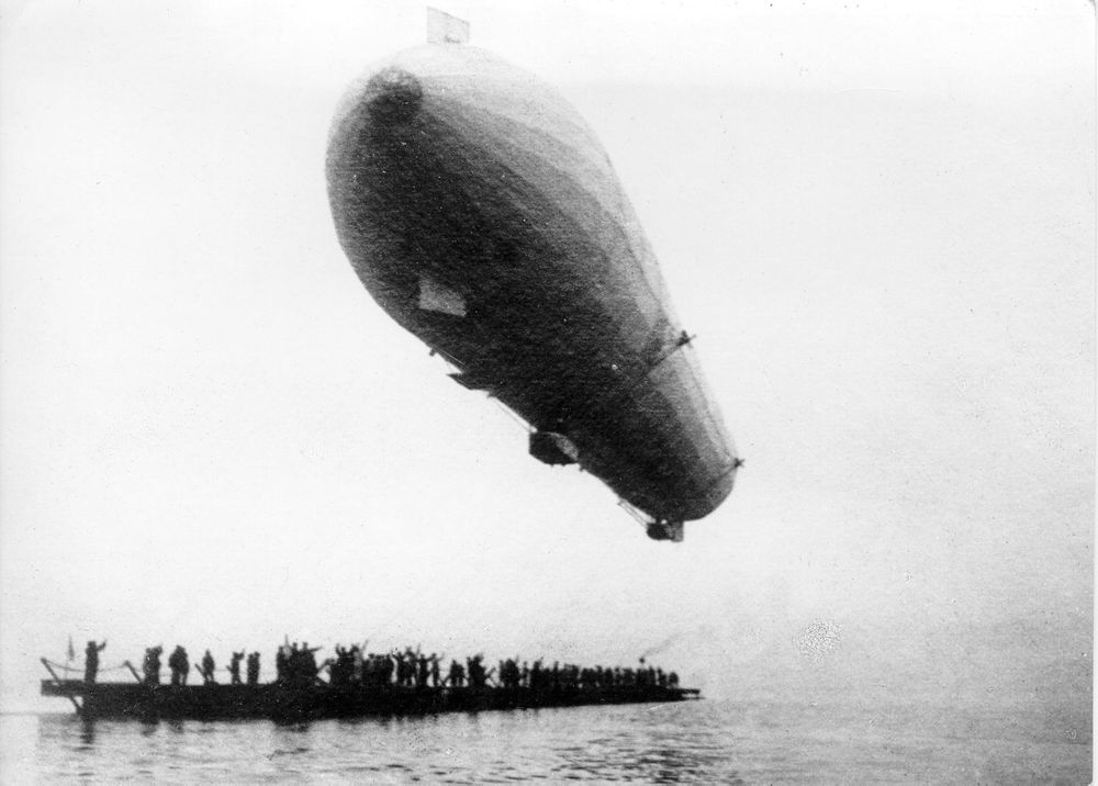 Der fahrende Zeppelin entfernt sich vom Ponton. Auf dem vollbesetzten Ponton jubeln die Schaulustigen.