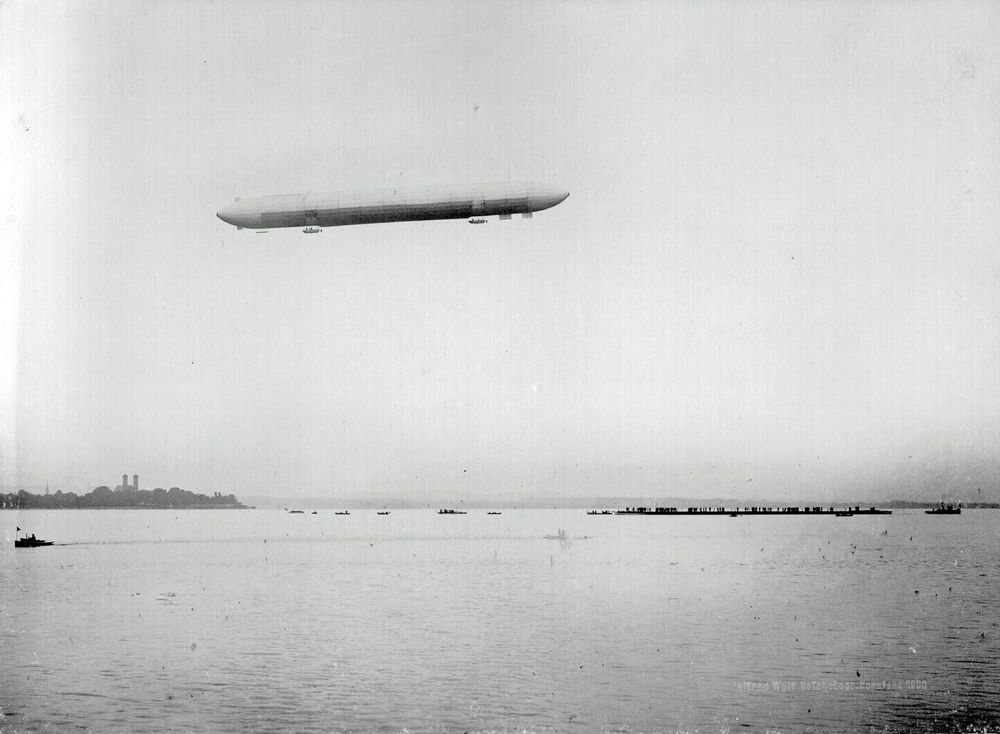 Der Zeppelin schwebt zwischen der Schloßkirche und dem vollbesetzten Ponton. Einige Boote mit Menschen darauf fahren über den See, um das Spektakel zu beobachten.