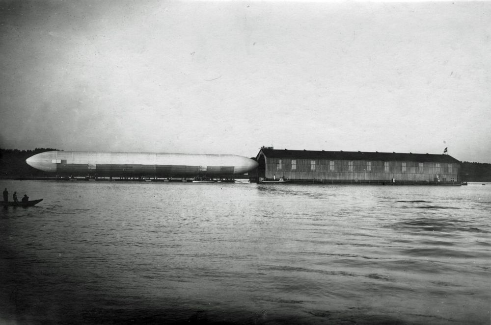 Der Zeppelin wurde gerade aus der Halle herausgefahren. Links fährt ein kleines Boot mit drei Personen.