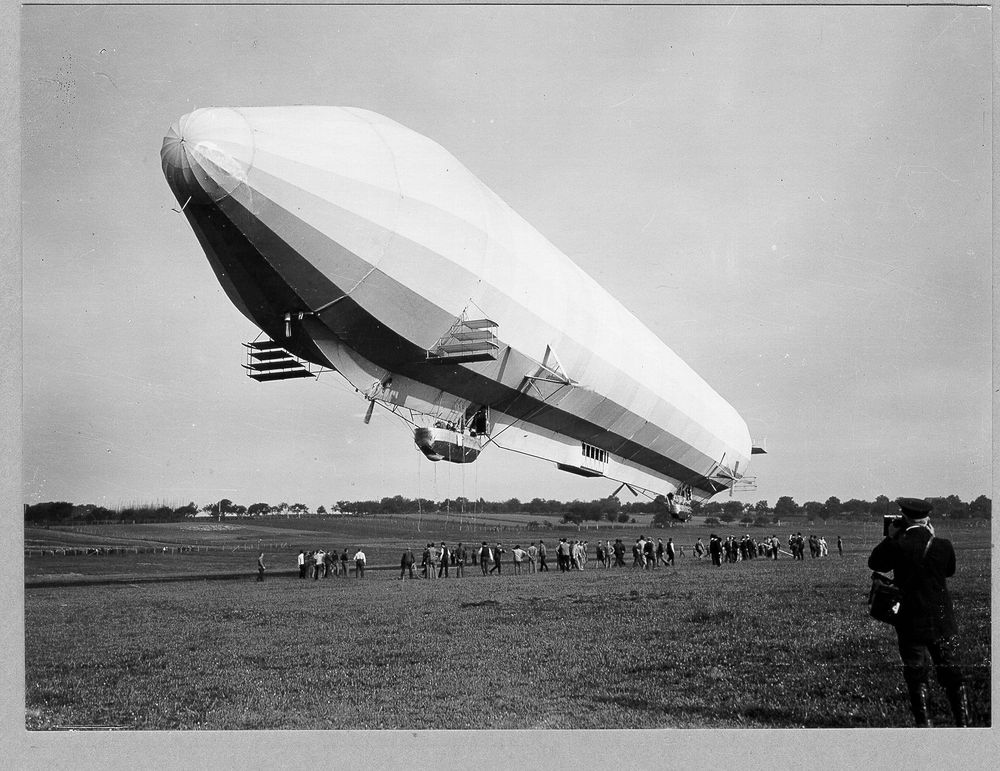 LZ 7 „Deutschland“ hebt gerade von einer Wiese ab. Im Hintergrund Bäume und Himmel, direkt unter dem Luftschiff die Haltemannschaft, bestehend aus etwa 100 Leuten.