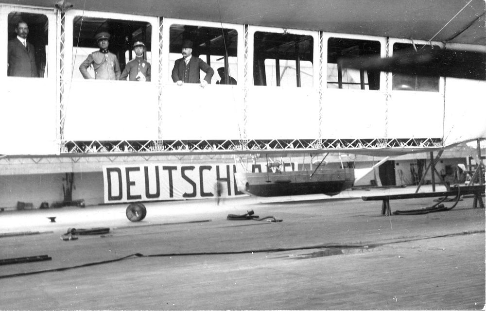 Seitenansicht der Passagiergondel des LZ 7 „Deutschland“ mit Menschen hinter den großen Panoramafenstern. Unterhalb des Luftschiffs ist eine Banderole angebracht, auf der „Deutschland“ steht.