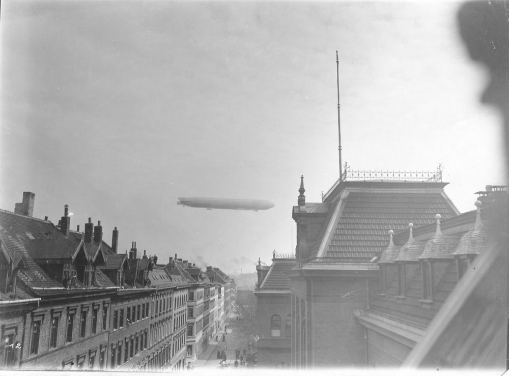 Die historische Aufnahme zeigt die Dächer einer Stadt. Im Hintergrund fliegt ein Luftschiff am Himmel.