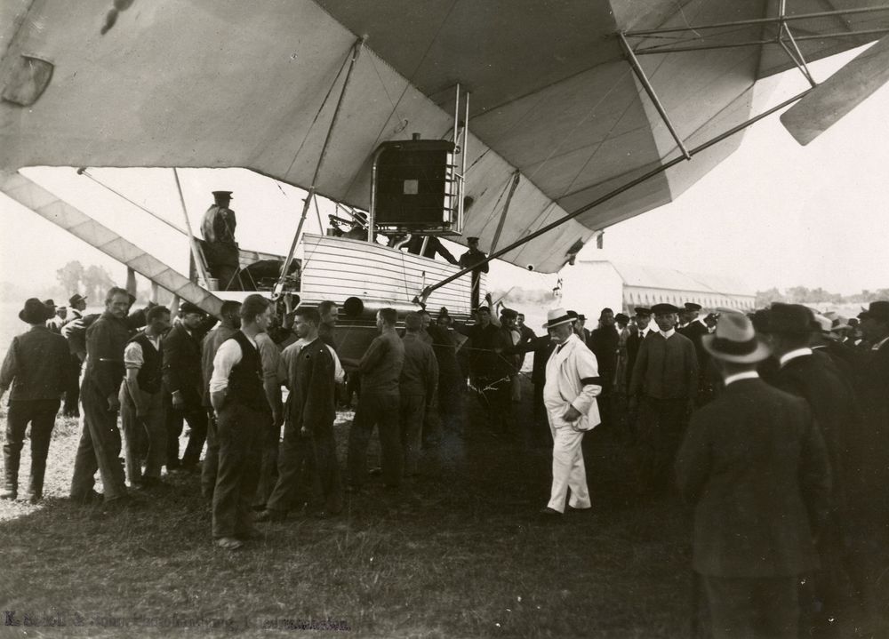 Die historische Aufnahme zeigt Graf Ferdinand von Zeppelin in einem weißen Anzug außerhalb einer Luftschiffgondel stehen. Um ihn herum befinden sich weitere Personen, darunter auch aus der Haltemannschafft.