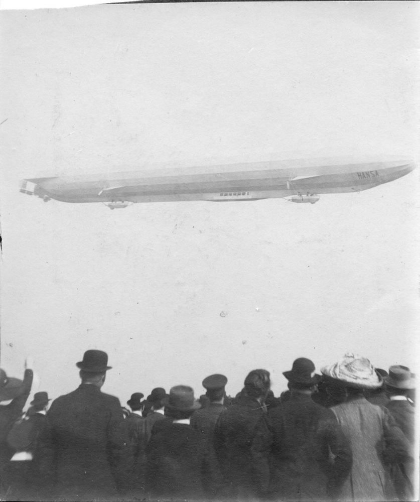 Die historische Aufnahme zeigt einen Zeppelin am Himmel, darunter betrachtet eine große Menschenmenge das Luftschiff am Himmel.
