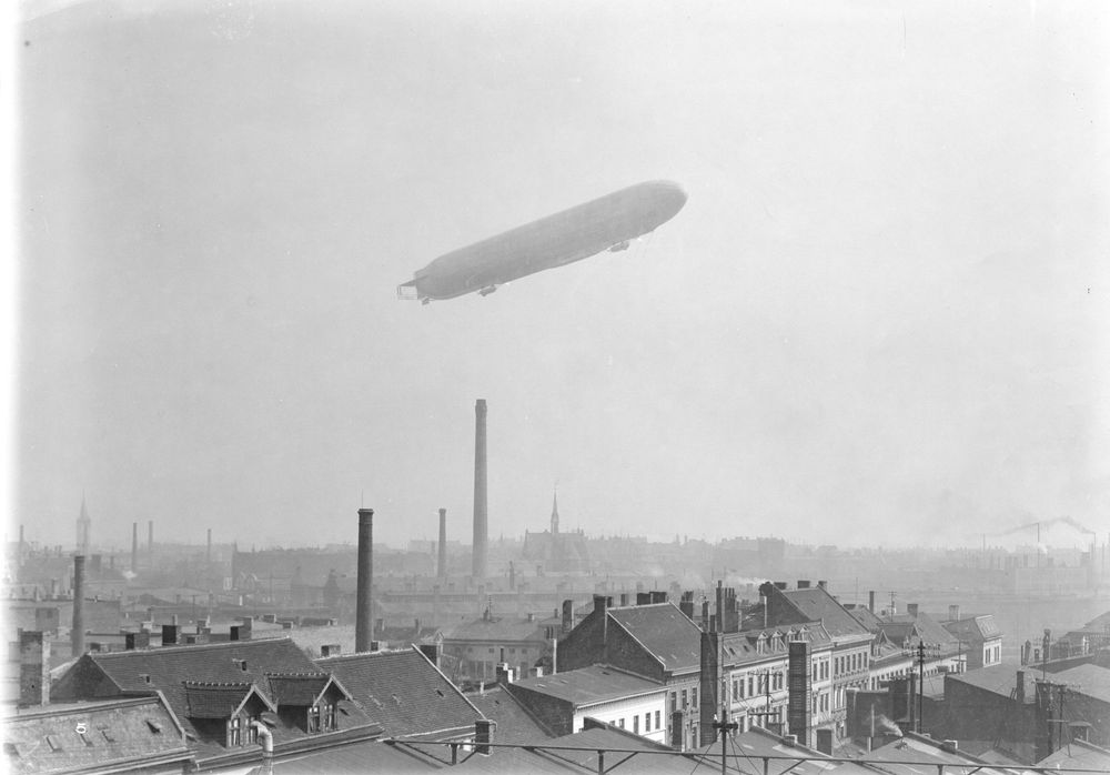 Die historische Aufnahme zeigt die Dächer einer Stadt. Im Hintergrund fliegt ein Luftschiff am Himmel.