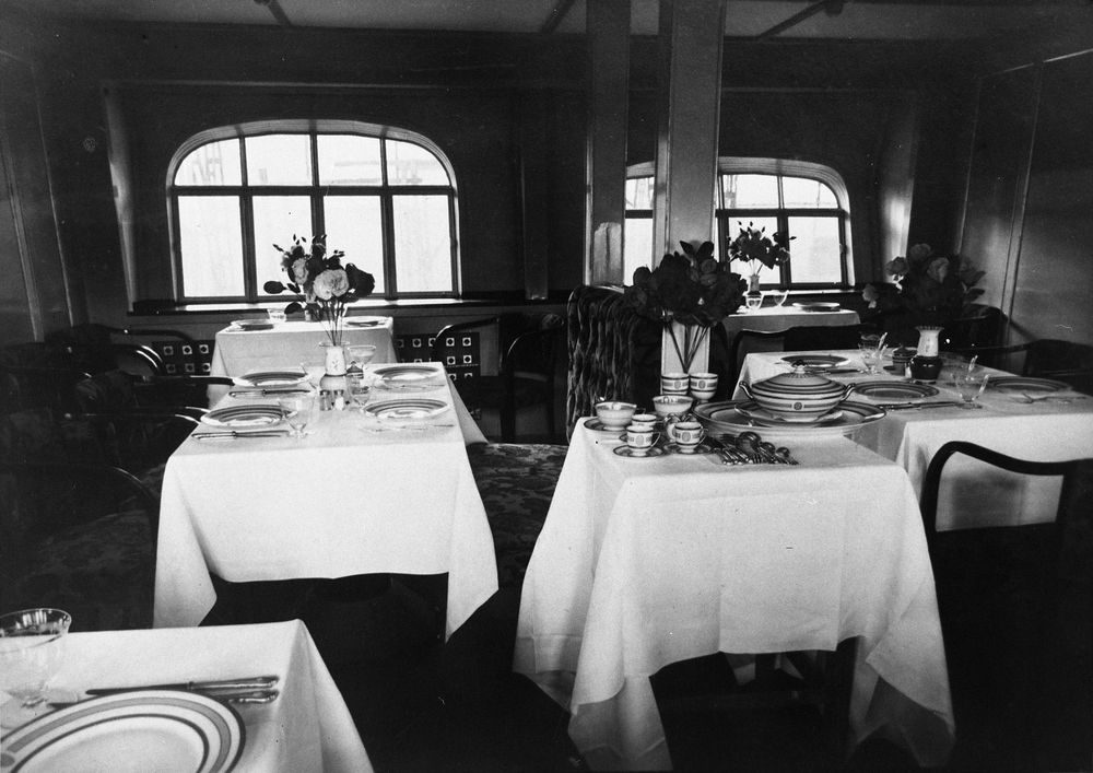 Die historische Aufnahme zeigt die Inneneinrichtung eines Aufenthaltsraum im Zeppelin. Im Raum sind mehrere Tische und Stühle verteilt. Die Tische sind zum Essen gedeckt.