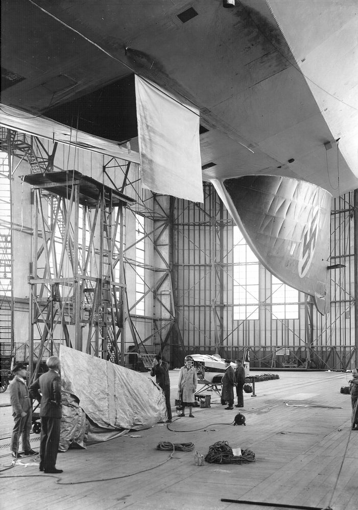 Vom Schiffsrumpf aus gesehen der steile Blick auf das untere Seitenleitwerk. Dort ist vollflächig das Hakenkreuz aufgebracht. Unter dem Zeppelin stehen Menschen.