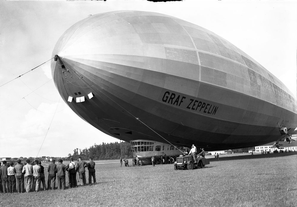 Die historische Aufnahme zeigt einen Zeppelin bei der Landung, der von einer Haltemannschaft bestehend aus einigen Männern und einem Raupenschlepper zu Boden gezogen wird.