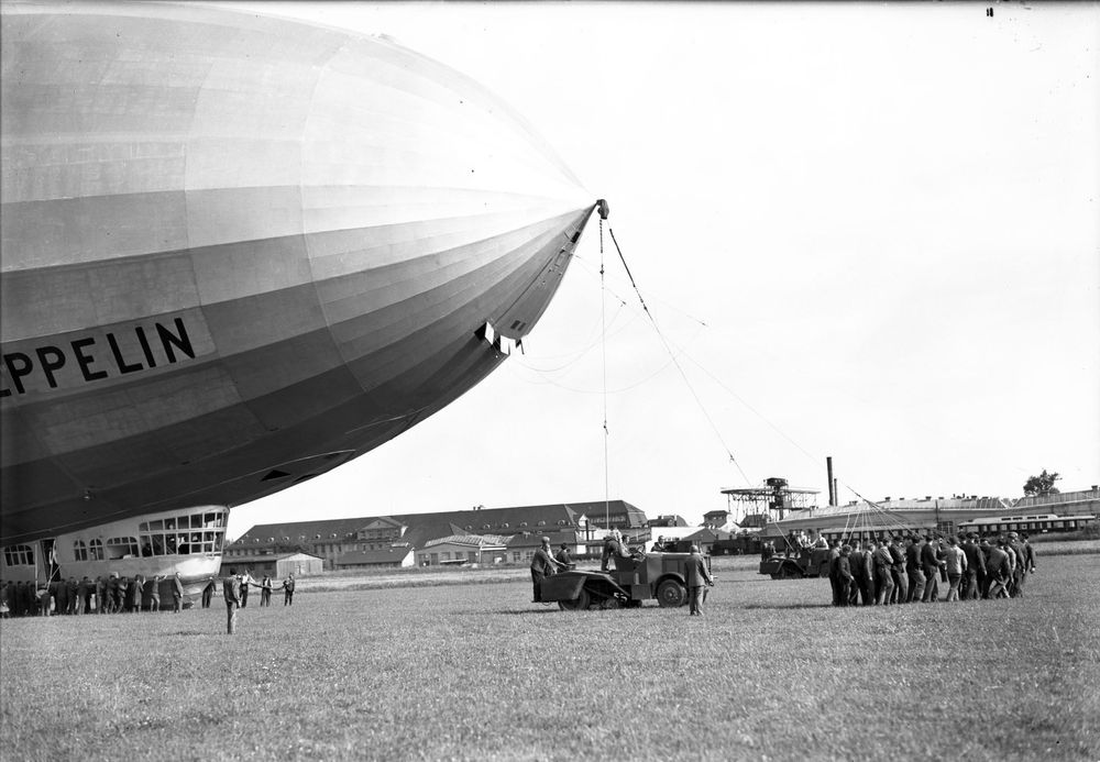 Die historische Aufnahme zeigt einen Zeppelin bei der Landung, der von einer Haltemannschaft bestehend aus einigen Männern und einem Raupenschlepper zu Boden gezogen wird.