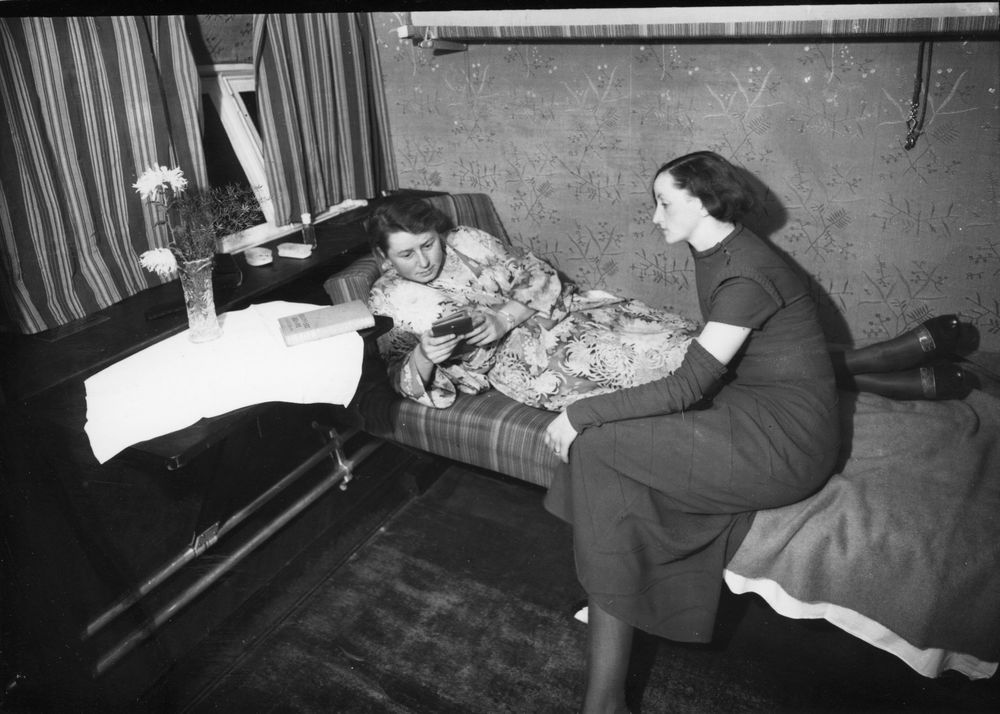 Die historische Aufnahme zeigt zwei Luftschiff-Passagierinnen, die sich in einer Kabine befinden. Eine der Frauen liegt auf dem Bett, die andere Frau sitzt daneben.