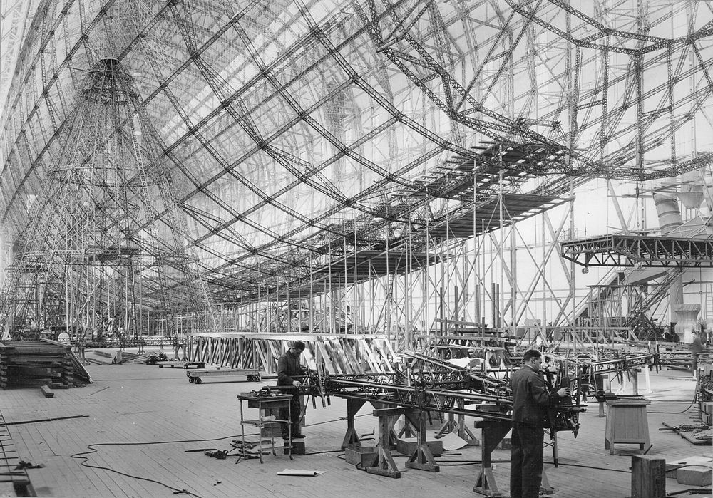 Ein riesiges Luftschiff befindet sich im Bau. Arbeiter stehen an Metallgerüsten und montieren Teile des massiven Rahmens. Die Struktur besteht aus zahlreichen Verstrebungen, die das Gerüst des Zeppelins formen.