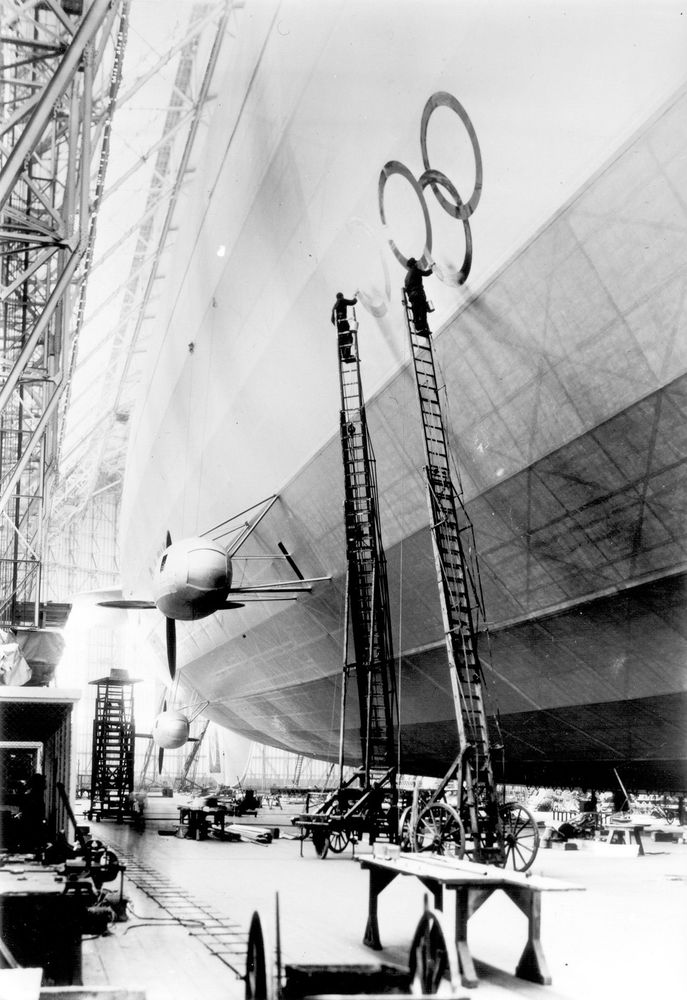 Schwarz-Weiß-Foto der „Hindenburg“ in der Halle. Man sieht die Hülle und zwei Propeller des Luftschiffs. Gerade werden die Olympiaringe durch zwei Arbeiter auf Drehleitern aufgemalt. Auf dem Boden der Halle liegt verschiedenes Handwerksgerät.