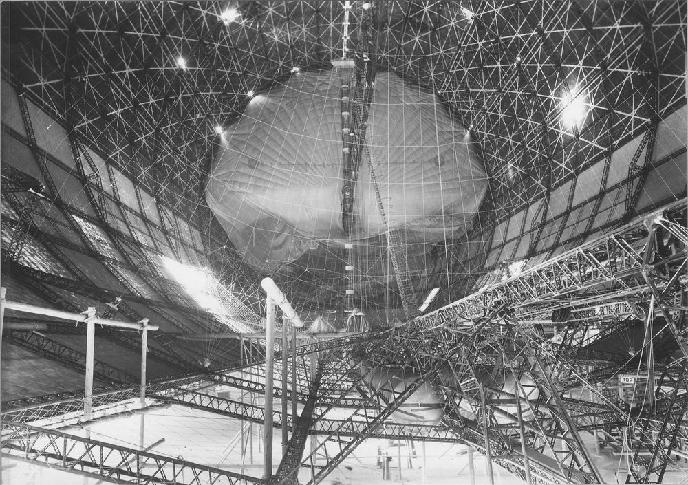 Eine riesige Luftschiffhalle mit einer teilweise fertiggestellten Zeppelin-Struktur. Das komplexe Metallgerüst des Zeppelins ist deutlich sichtbar, während die Hülle erst teilweise angebracht wurde.