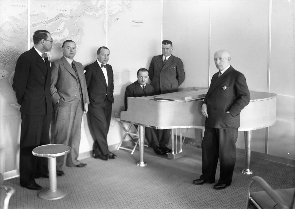 Die historische Aufnahme zeigt eine Gruppe von Männern, die um ein Klavier stehen, an dem ein weiterer Mann sitzt.