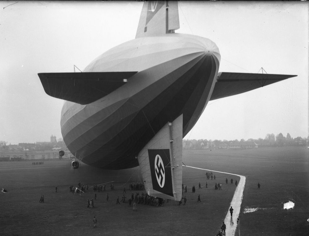 Der Zeppelin in der leicht verschobenen Heckansicht. Das obere Ende des Luftschiffs passt nicht mehr ganz aufs schwarz-weiß Foto. Auf den Seitenleitwerken sind die Hakenkreuzfahnen aufgemalt zu sehen. Unter dem Zeppelin befinden sich einige Menschen.