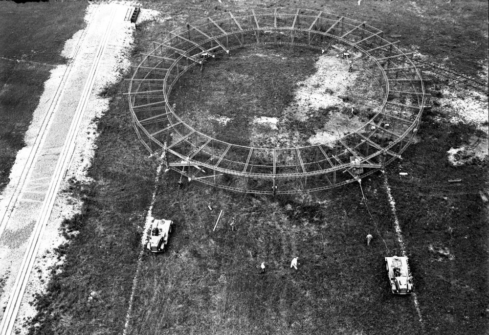 Die historische Aufnahme zeigt ein Feld aus der Vogelperspektive, auf dem zwei Raupenschlepper einen Gerippe-Ring eines Zeppelins ziehen.