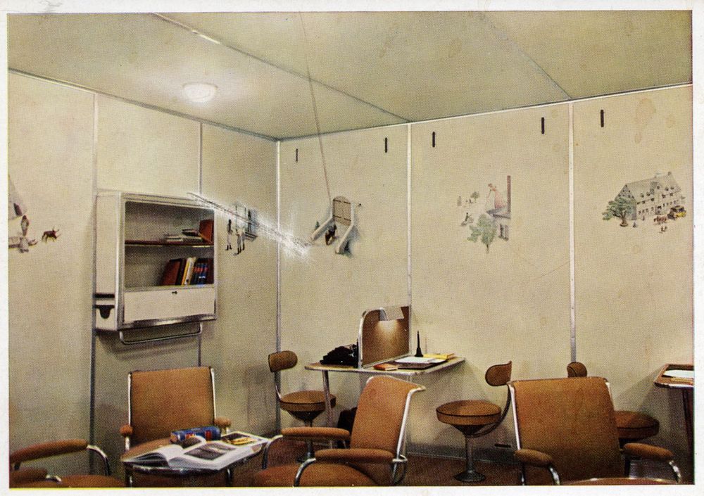 Farbige Postkarte mit Blick auf das Post-, Schreib- und Lesezimmer von LZ 129. Umlaufend sind Bemalungen erkennbar, links ein Bücherschrank, im Vordergrund Stühle und Tische. Das Bild weist über der kolonialen Darstellung einen tiefen Kratzer auf. 