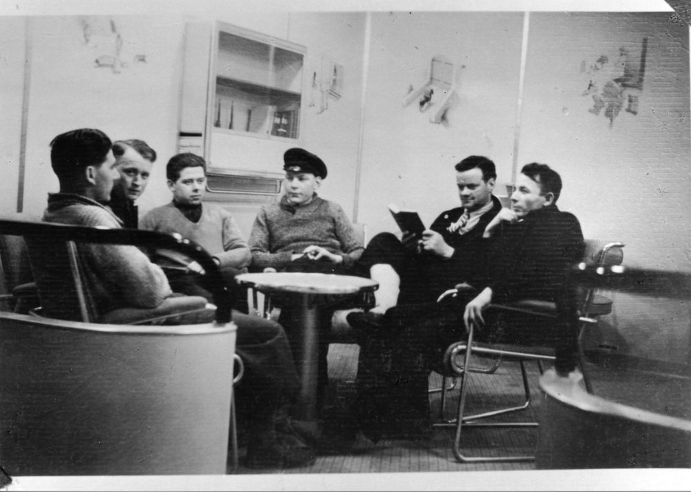 Eine Gruppe junger Männer im Post- Schreib- und Lesezimmer sitzt am Tisch und unterhält sich. Im Hintergrund der Bücherschrank und die Wandmalereien. 